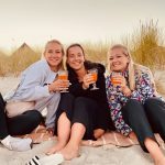 3 Teilnehmerinnen sitzen mit einem Drink am Strand in der Abenddämmerung